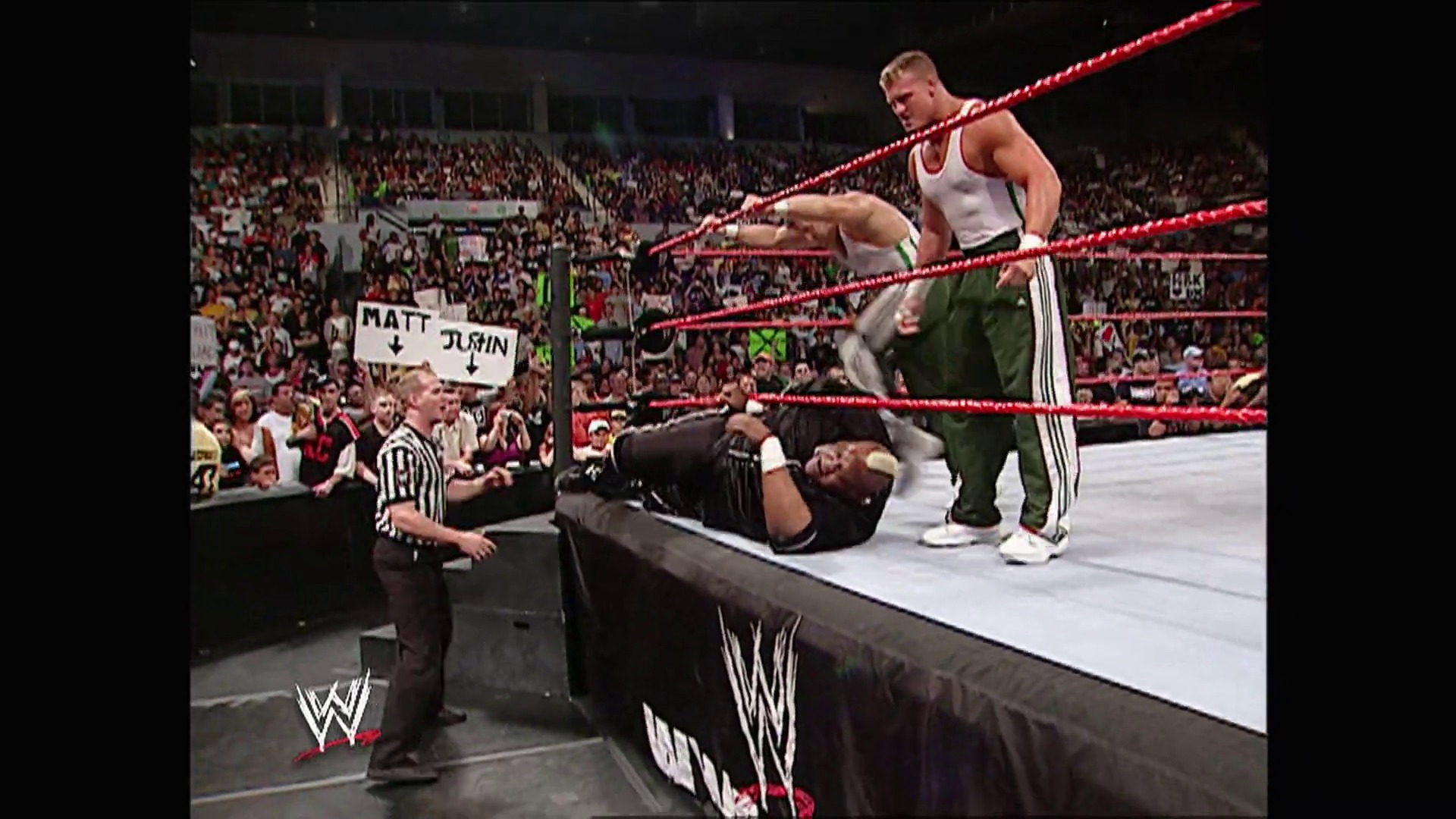 A frame from WWE Raw (June 19, 2006) at 23:27. A WWE wrestler in a white tank top stands over a fallen opponent on the red-roped ring, while a referee watches nearby. A packed arena crowd fills the background, with a visible fan sign reading "MATT" and "JJJOHN" as the WWE logo appears at the bottom of the screen.