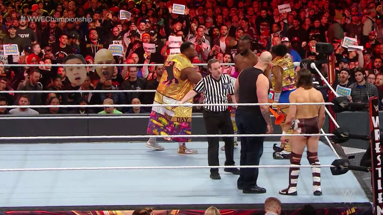 A frame from WrestleMania 35 (2019) at 2:17:12. A WWE wrestler in gold and purple attire stands in the ring with a referee during WrestleMania 35, facing an opponent in burgundy and white. The packed crowd in red fills the arena in the background, with visible WWE Championship graphics displayed overhead.