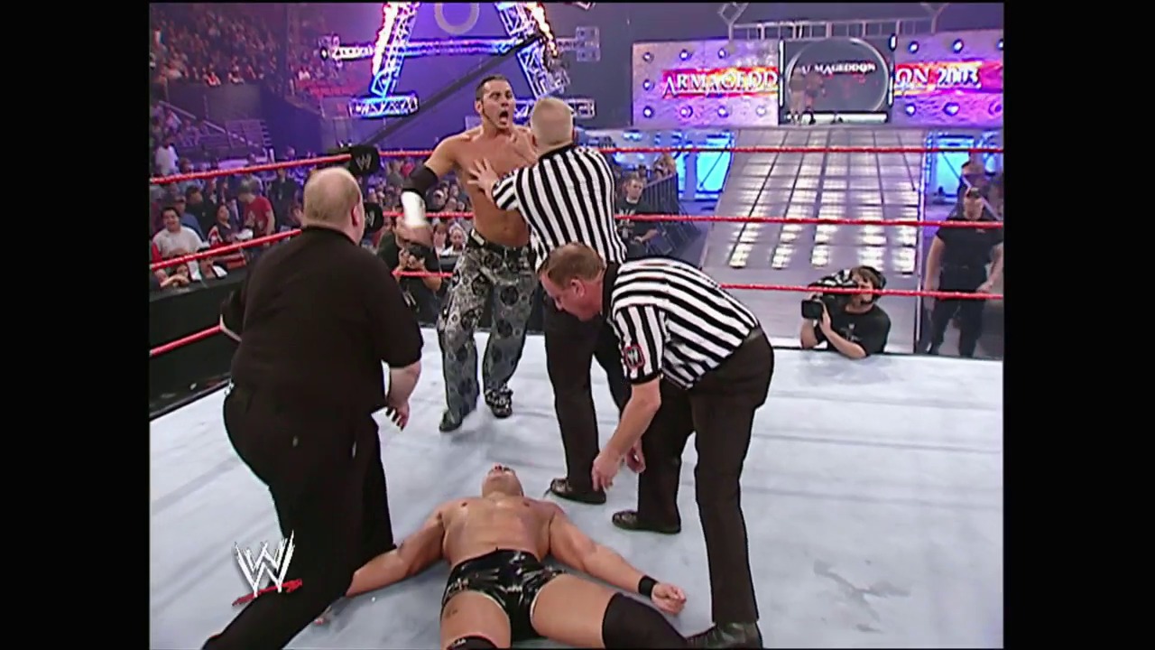 A frame from WWE Armageddon (2003) at 1:27:46. A WWE wrestling match in progress at Armageddon 2003, with one wrestler lying on the mat while another stands nearby. Referees in striped shirts oversee the action as the crowd watches from the arena seats, with purple lighting and WWE graphics visible in the background.