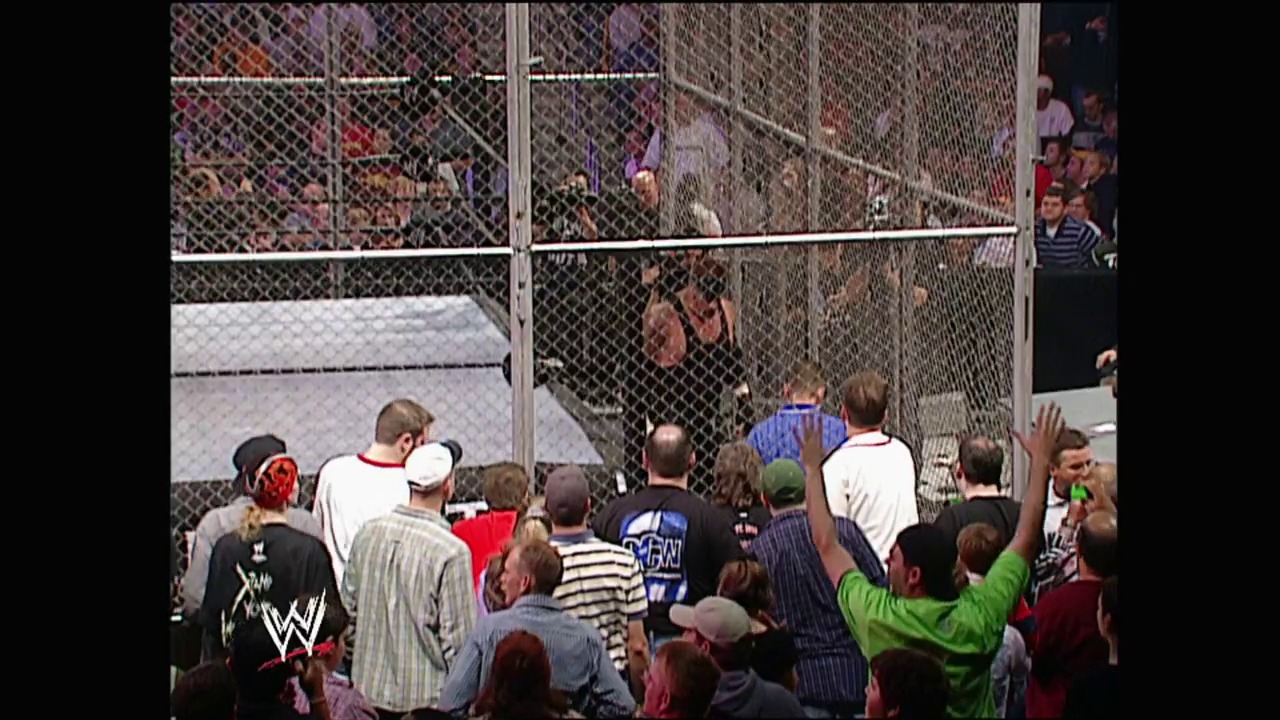 A frame from WWE No Mercy (2002) at 2:21:37. A crowd of spectators gathered around a steel cage structure during a WWE wrestling match, with fans in various colored shirts visible both inside and outside the enclosure, appearing to be an intense moment from the event.