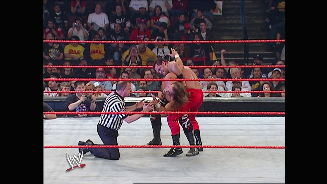 A frame from WWE Raw (February 16, 2004) at 1:20:00. A WWE referee in black and white stripes kneels in the red-roped ring, observing two wrestlers in combat. The crowd fills the arena stands in the background, with the WWE logo visible in the lower left corner.