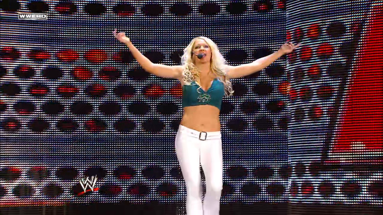 A frame from WWE Raw (January 5, 2009) at 53:37. A blonde female wrestler stands on stage with both arms raised in celebration, wearing a teal sports bra and white pants with a belt. She performs in front of a large LED screen displaying the WWE logo and red/black geometric patterns.