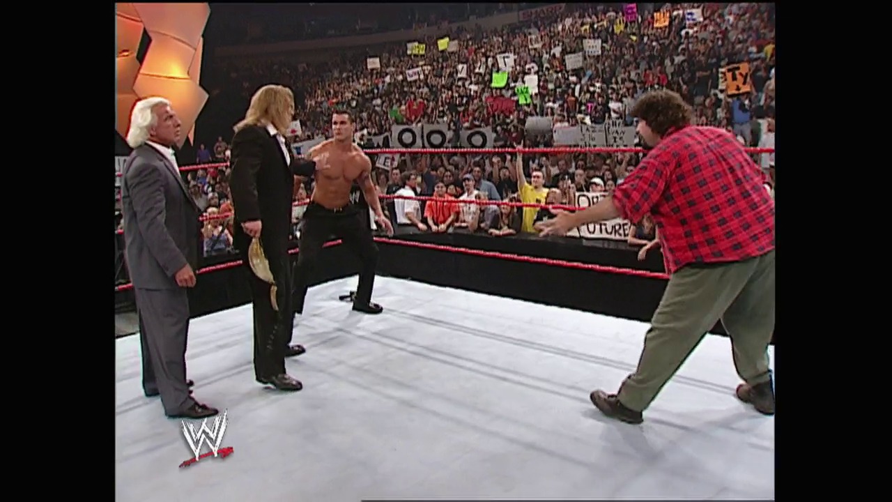 A frame from WWE Raw (June 16, 2003) at 13:46. A WWE Raw wrestling scene features multiple people in and around the ring, including an shirtless wrestler, suited officials, and a man in a red plaid shirt. A packed stadium crowd fills the background behind the red ropes, with various signs visible throughout the arena.