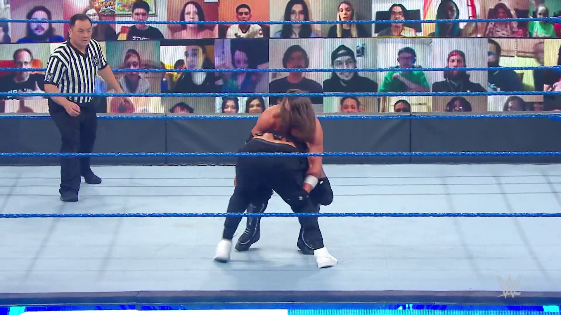 A frame from WWE SmackDown (October 2, 2020) at 24:21. A WWE wrestler in black attire crouches in the ring while a referee in striped shirt officiates. Virtual audience screens line the background, reflecting the pandemic-era broadcast setup.