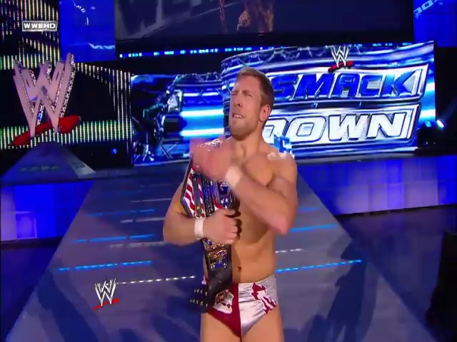 A frame from WWE SmackDown (October 29, 2010) at 24:14. A shirtless WWE wrestler stands in the ring wearing a patriotic-themed singlet, holding a championship belt. The blue-lit SmackDown stage is visible in the background with WWE branding and graphics.