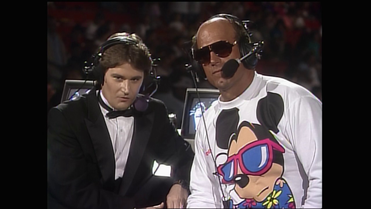 A frame from Royal Rumble (1990) at 1:32:58. Two commentators wearing headsets sit at the broadcast table during the event. The man on the right wears sunglasses and a white shirt with a cartoon character graphic, while the man on the left is dressed in formal attire. A blurred crowd is visible in the background.