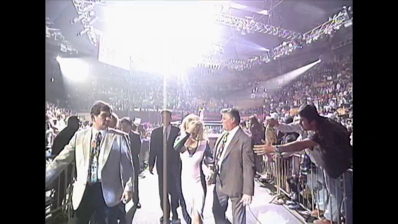 A frame from Royal Rumble (1995) at 2:42:15. A large indoor arena filled with spectators during a wrestling event, with several formally dressed individuals standing on the arena floor beneath bright overhead lighting. The crowd is visible in the background bleachers, and the scene captures the ceremonial atmosphere of a major wrestling production.