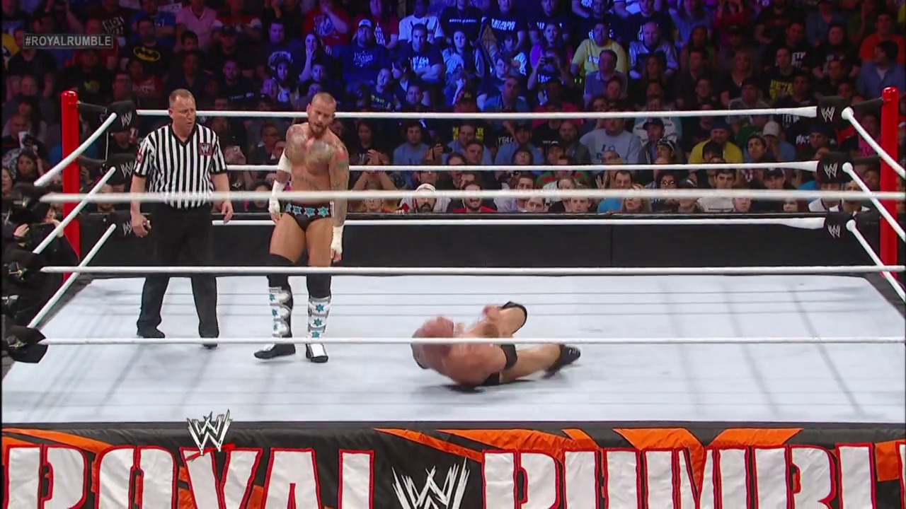 A frame from Royal Rumble (2013) at 2:22:42. A WWE wrestler lies on the mat after being knocked down during a Royal Rumble match, while an opponent stands nearby and a referee oversees the action. The packed arena crowd watches intently from the stands surrounding the white ring with red corner posts.