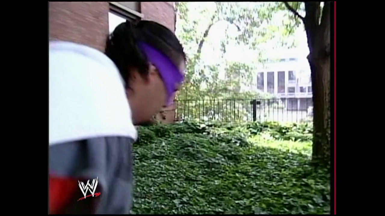 A frame from WWE Raw (August 18, 2003) at 53:23. A wrestler wearing a white shirt and purple bandana is shown in profile at an outdoor location with hedges and buildings visible in the background. The WWE logo appears in the lower left corner of the frame.