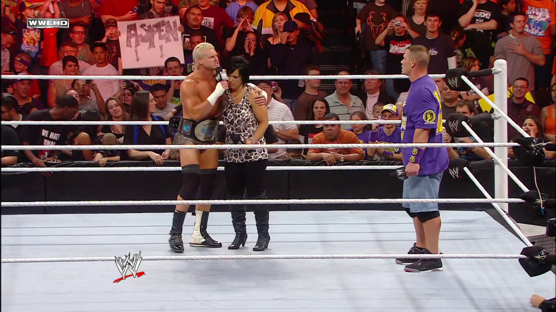 A frame from WWE Raw (December 20, 2010) at 48:05. A WWE wrestler with blonde hair holds a microphone in the ring while a woman in a patterned dress stands nearby, with another wrestler in purple visible at ringside. The packed crowd watches from the stands as the WWE logo appears on the white canvas.