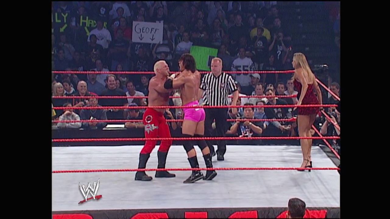 A frame from WWE Raw (June 2, 2003) at 26:26. Two male wrestlers engage in combat inside a WWE ring bordered by red ropes, while a referee in black and white stripes oversees the match. A blonde female wrestler stands nearby as the crowd watches from the arena seating in the background.