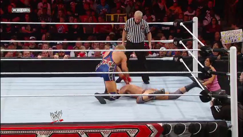 A frame from WWE Raw (November 28, 2011) at 1:03:34. A WWE wrestler in blue and red attire lies on the mat while an opponent stands nearby, with a referee in striped shirt overseeing the match. The packed crowd fills the arena background as the action unfolds in the white-roped ring.