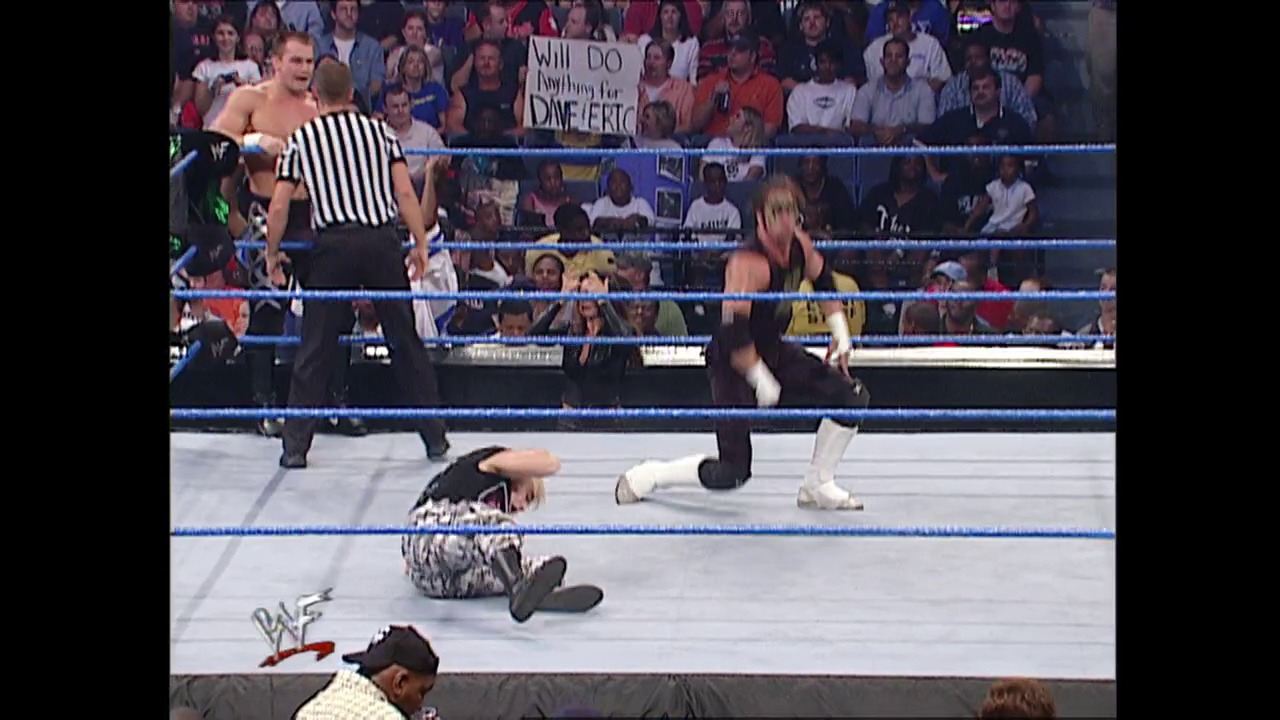 A frame from WWE SmackDown (September 20, 2001) at 26:35. A WWE SmackDown match in progress features wrestlers in and around the ring, with a referee in a striped shirt overseeing the action. The crowd fills the arena in the background, and a fan's sign reading "Will DO anything for DAVE FERTO" is visible above the ring.