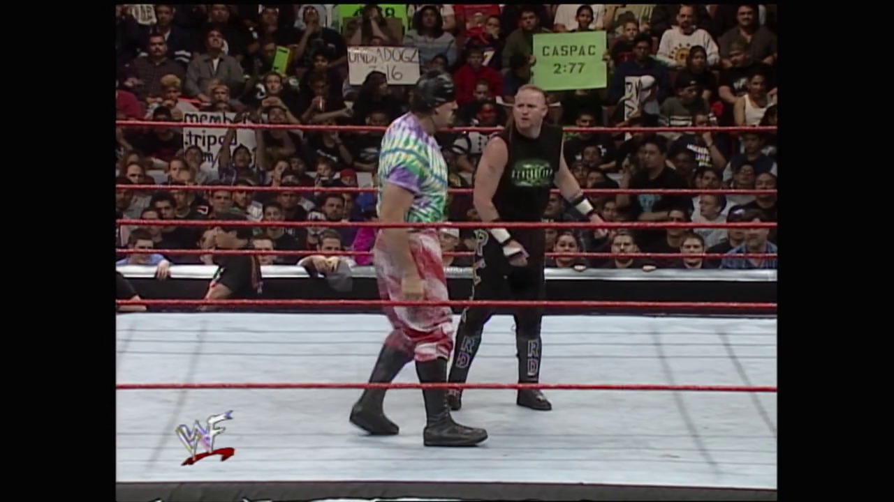 A frame from WWF Royal Rumble: No Chance in Hell (1999) at 2:03:04. Two wrestlers face off in a WWF ring during a match, with one in colorful neon attire and the other in black. A packed crowd fills the arena stands behind the red-roped ring, with the WAF logo visible in the corner.