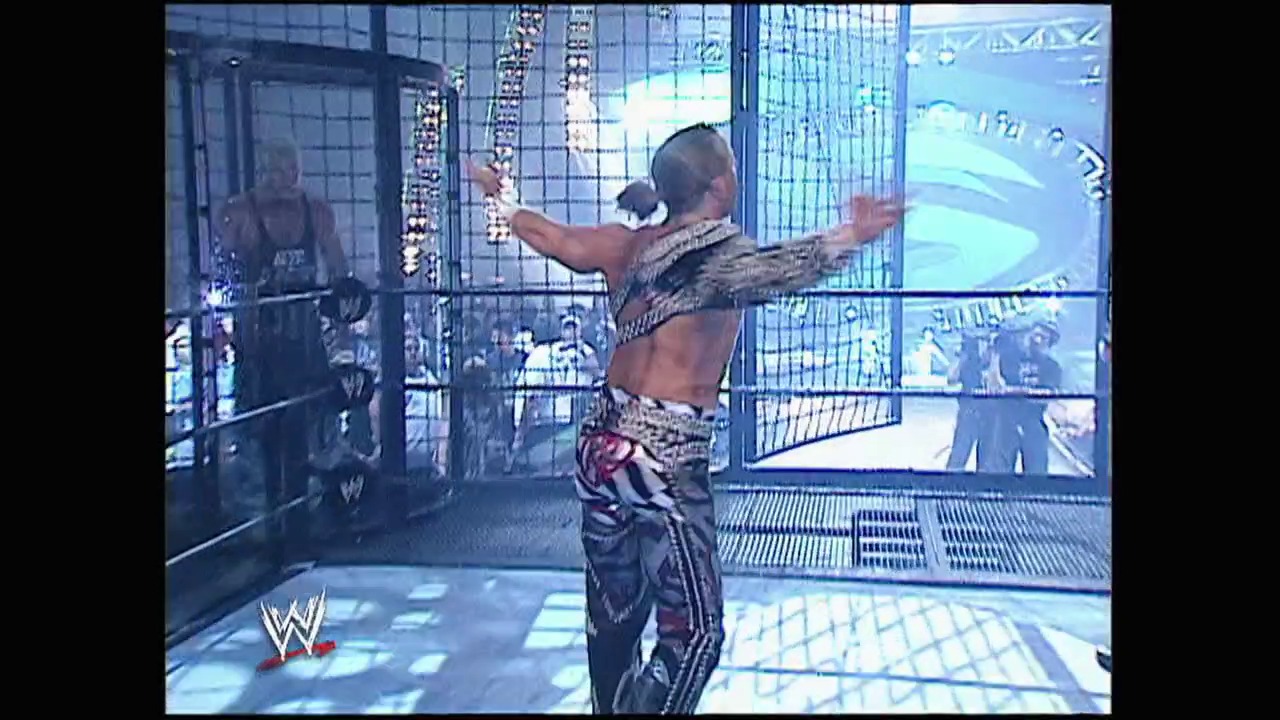 A frame from Summerslam (2003) at 2:20:07. A shirtless wrestler in patterned pants and headphones stands with arms raised in a modern wrestling ring with glass panels, while spectators and arena infrastructure are visible in the background during a WWE event.