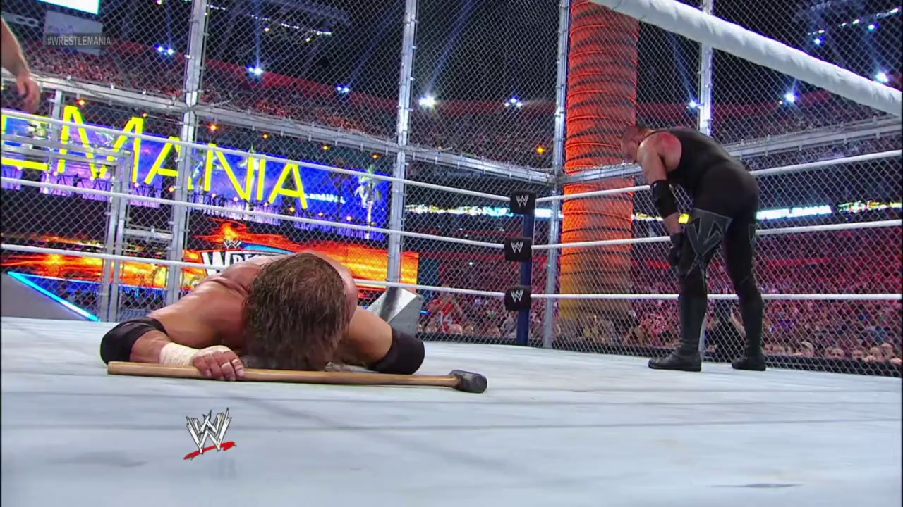 A frame from WrestleMania XXVIII (2012) at 1:38:53. A wrestler lies prone on the steel cage floor holding a weapon, while another competitor stands nearby in a Steel Cage match at WrestleMania XXVIII, with the illuminated arena and packed crowd visible in the background.