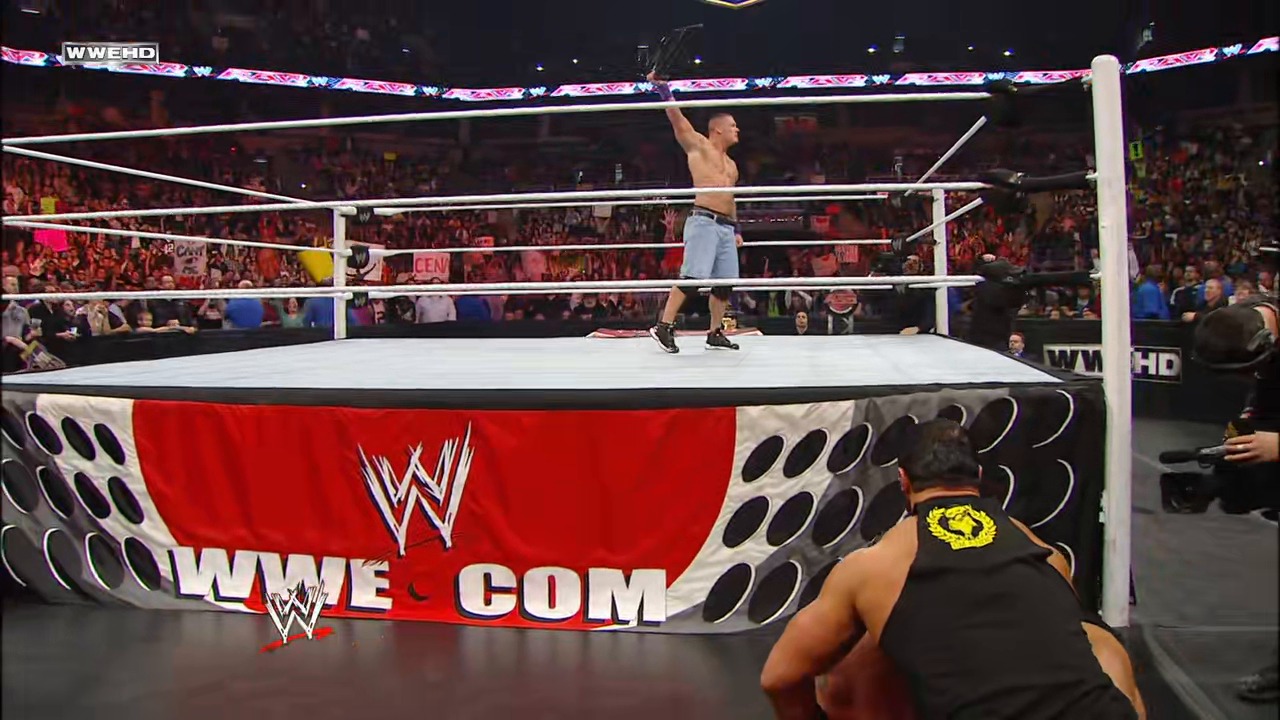 A frame from WWE Raw (February 7, 2011) at 1:27:35. A shirtless wrestler stands victoriously on the white WWE ring ropes while an opponent approaches from below. The arena is packed with enthusiastic fans, and the red WWE branded barrier is visible in the foreground with the "WWE.COM" logo displayed.