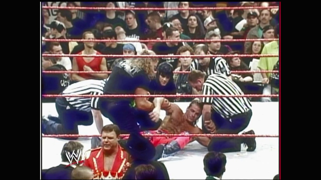 A frame from WWE Raw (January 19, 2004) at 26:02. A WWE wrestler in a red and gold outfit lies on the mat while opponents in dark clothing grapple nearby, with a referee in striped shirt overseeing the action. The crowd watches from the stands behind the red-roped ring, with the WWE logo visible in the corner.