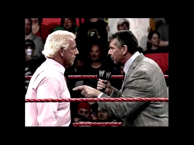 A frame from WWE Raw (January 7, 2008) at 0:27. Two men in business attire engage in a confrontation inside a WWE wrestling ring surrounded by a live crowd. One wears a pink shirt while the other sports a gray suit, both appearing to be in an intense discussion near the red ropes.