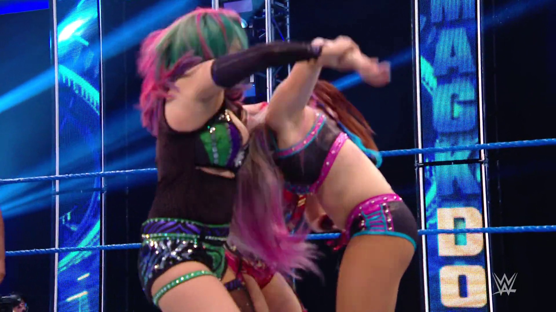 A frame from WWE SmackDown (April 10, 2020) at 12:37. A WWE wrestler with pink and green hair performs in the ring, holding a microphone while striking an energetic pose. The wrestling ring is surrounded by LED screens displaying blue graphics and the WWE logo, with blue rope visible in the foreground.