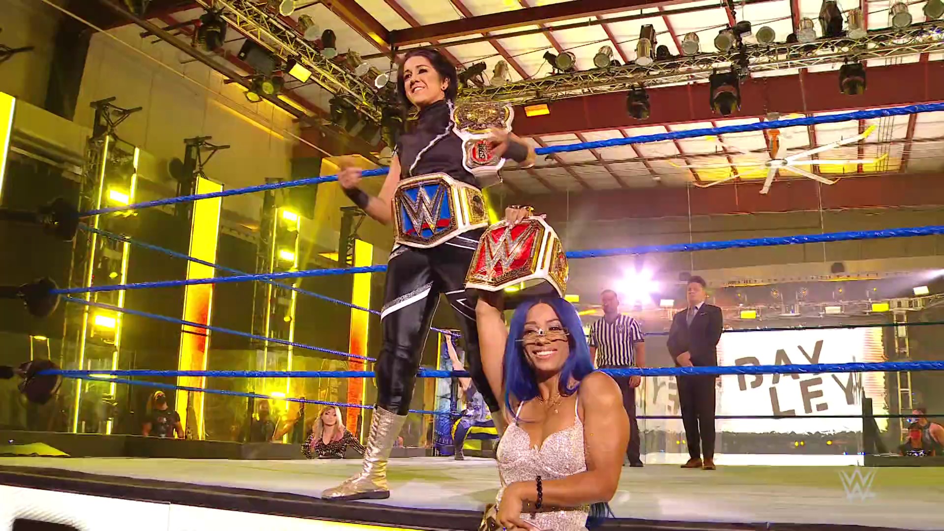 A frame from WWE SmackDown (July 31, 2020) at 1:12:34. # WWE SmackDown Ring Scene

A female wrestler stands triumphantly in the blue WWE ring holding championship belts, while another performer with blue hair smiles in the foreground. The venue features bright yellow and red lighting, with officials and spectators visible in the background.
