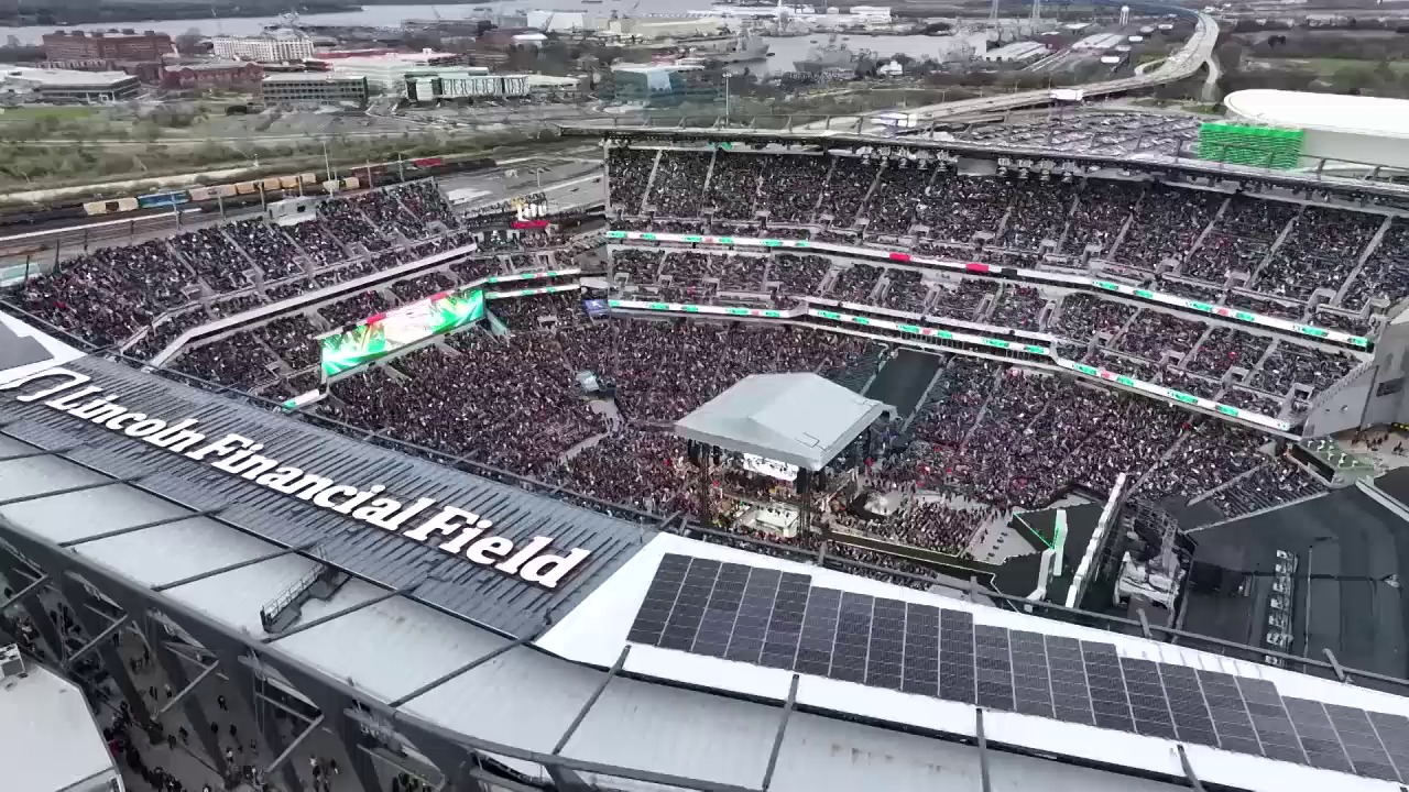 A frame from WWE WrestleMania 40 Saturday (2024) at 1:00. # WrestleMania 40 Stadium View

A packed stadium filled with thousands of enthusiastic fans surrounds a wrestling ring with a stage setup in the center. Solar panels are visible on the stadium roof, while green digital displays and signage frame the massive crowd gathered for the WWE event.