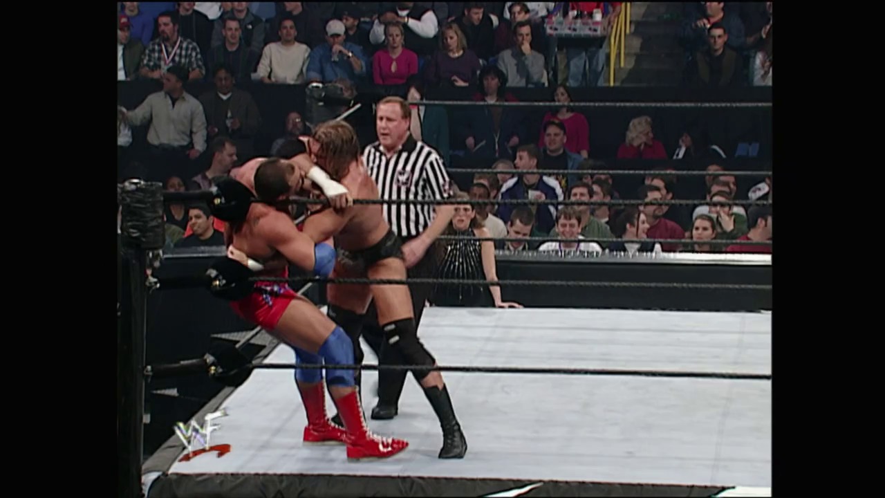 A frame from Royal Rumble (2001) at 1:13:21. Two wrestlers grapple in the ring during an intense match, with a referee in striped shirt overseeing the action. The crowd fills the arena bleachers in the background while the WWF logo is visible in the corner.