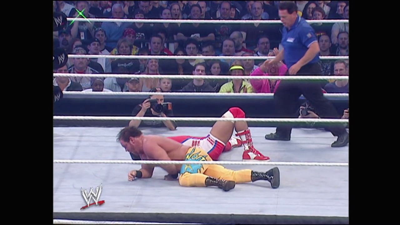 A frame from WrestleMania 23 (2007) at 1:03:59. Two WWE wrestlers are down on the mat during an intense match, with one in red and white attire and another in yellow and blue. The crowd watches from the stands while a referee in blue stands nearby in the professional wrestling ring.
