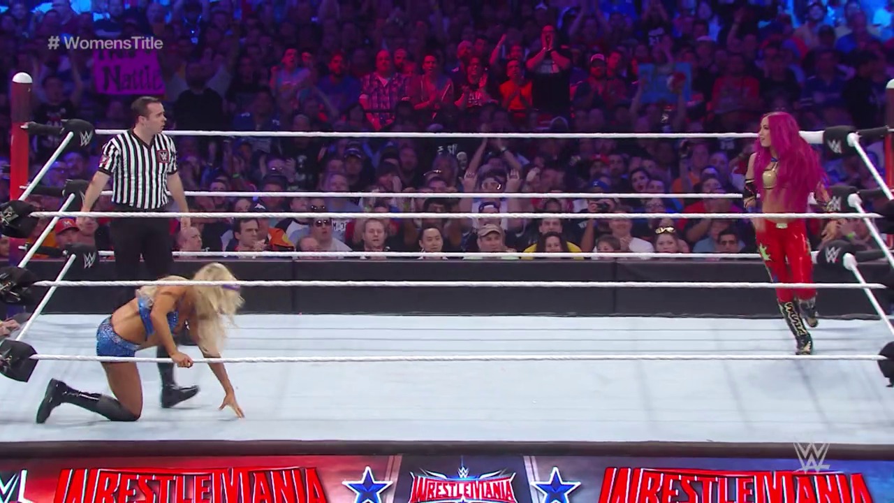 A frame from WrestleMania 32 (2016) at 2:04:34. A WWE Women's Title match at WrestleMania 32 features a blonde wrestler on her knees in the ring, a referee in striped shirt, and an opponent in red gear. The crowd is illuminated in purple and blue lighting in the background.