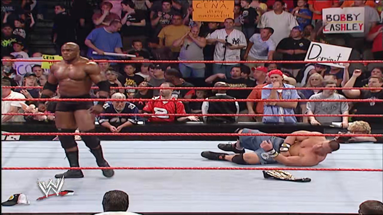 A frame from WWE Raw (July 2, 2007) at 1:27:59. A WWE wrestler stands over a fallen opponent on the mat during Raw. The crowd fills the arena with visible signs including "Bobby Lashley" and "Dominant," while the red ropes and WWE logo frame the action.
