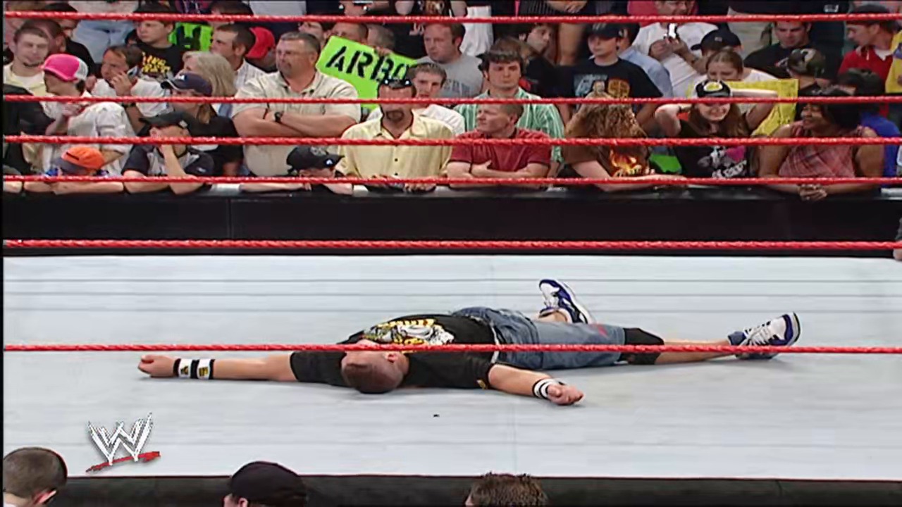 A frame from WWE Raw (May 21, 2007) at 46:24. A WWE wrestler lies motionless on the white mat in the red-roped ring, wearing black attire and a championship belt, while an engaged crowd watches from the stands above.