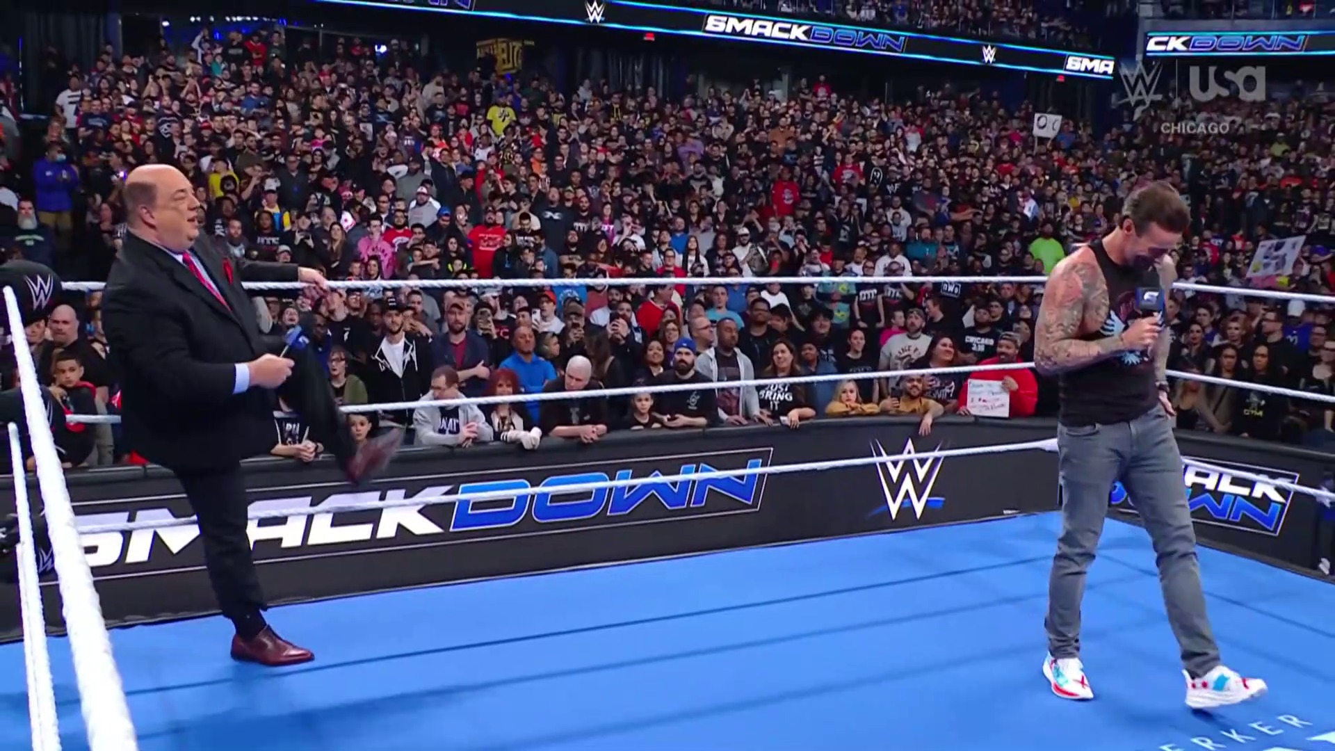A frame from WWE SmackDown (April 4, 2025) at 18:29. Two men stand in a WWE SmackDown ring before a packed Chicago crowd. One wears a black suit with a microphone on the left, while the other in casual attire holds a microphone on the right, with the blue mat and ropes visible behind them.
