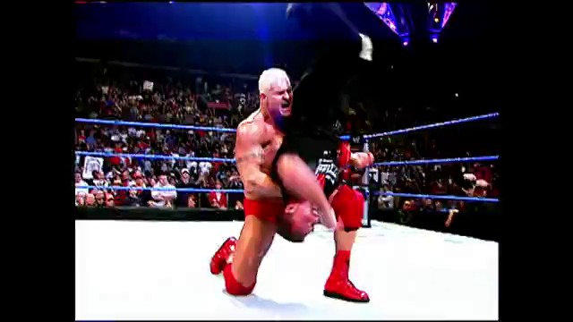 A frame from WWE SmackDown (March 25, 2005) at 0:45. A muscular wrestler in red attire performs in a WWE ring during SmackDown, with a packed crowd visible in the background and blue ring ropes framing the action.