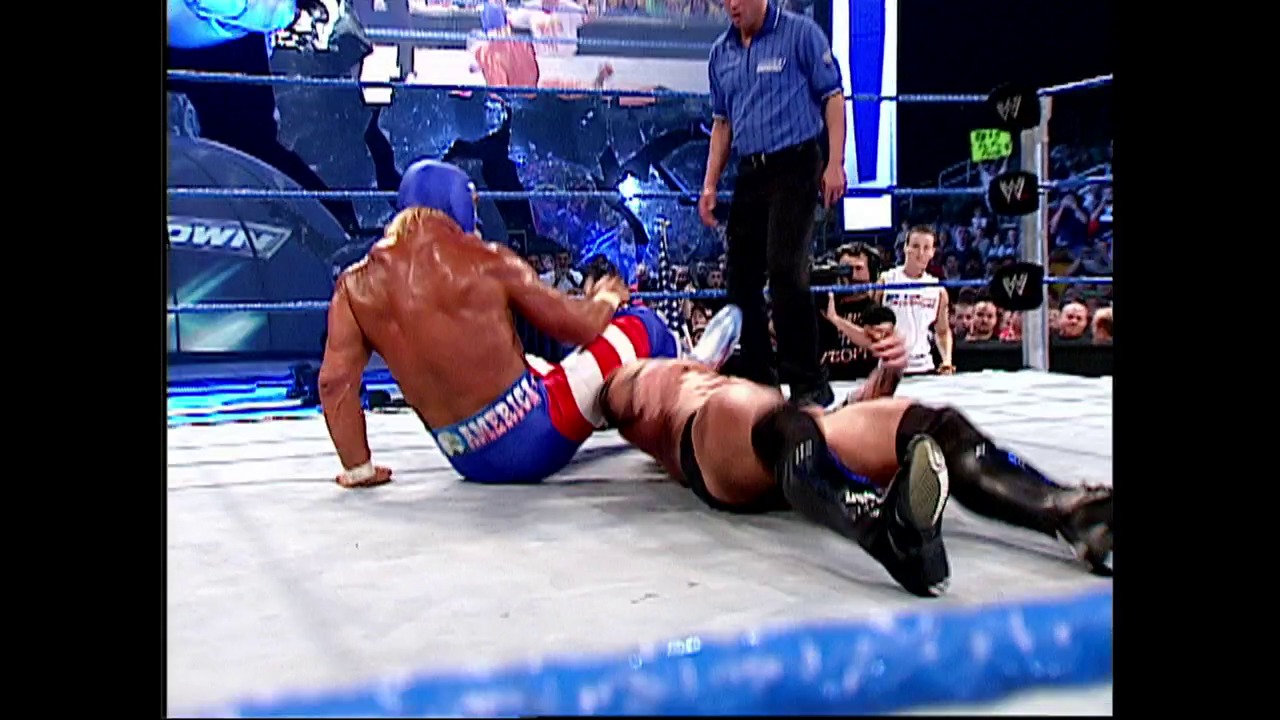 A frame from WWE SmackDown (May 29, 2003) at 0:54. A shirtless wrestler in blue and red attire is on the mat in a wrestling ring, with a referee in a blue shirt standing nearby and a crowd visible in the background. The WWE SmackDown branding and blue ring ropes are visible in the scene.