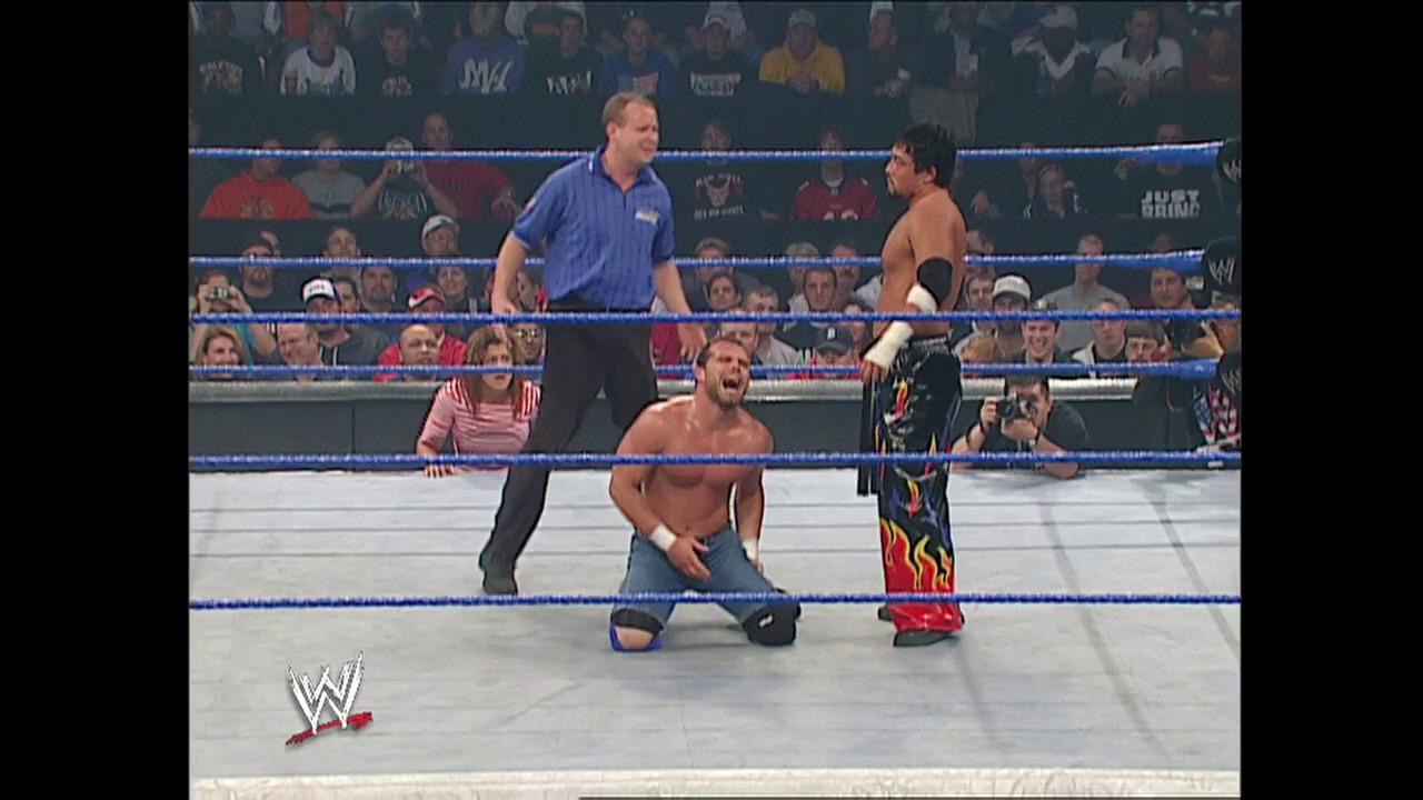 A frame from WWE SmackDown (October 16, 2003) at 37:49. A WWE wrestler in blue pants crouches in the ring facing an opponent in flame-patterned red and black attire, while a referee in a blue shirt oversees the match. The crowd fills the arena stands behind the blue-roped ring, with the WWE logo visible in the corner.