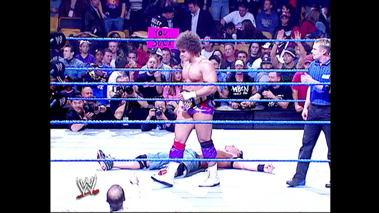 A frame from WWE SmackDown (October 7, 2004) at 1:26:07. A WWE SmackDown match features a standing wrestler in purple attire overlooking opponents on the canvas, with the blue-roped ring surrounded by an engaged crowd and ringside officials visible in the background.