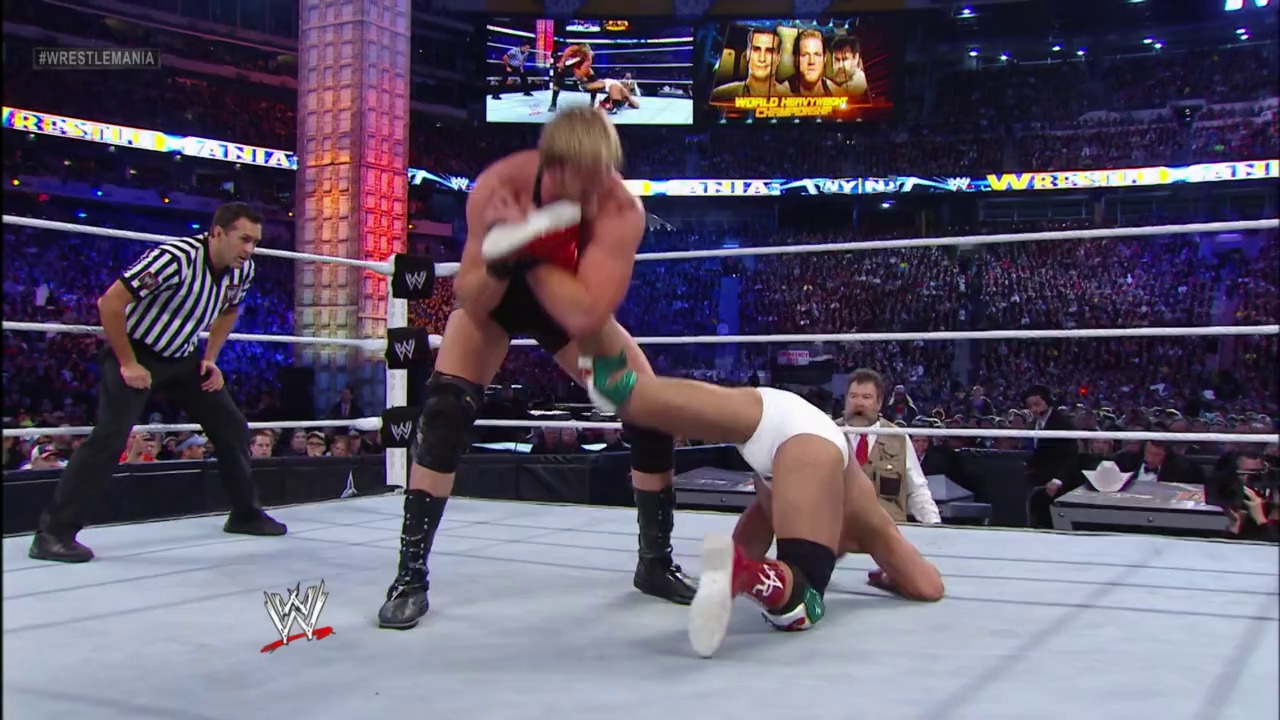 A frame from WrestleMania 29 (2013) at 1:37:05. A blonde wrestler performs a dynamic move in the WWE ring during WrestleMania 29, with a referee in striped shirt overseeing the match. The packed arena crowd is visible in the background alongside large video screens displaying the event.