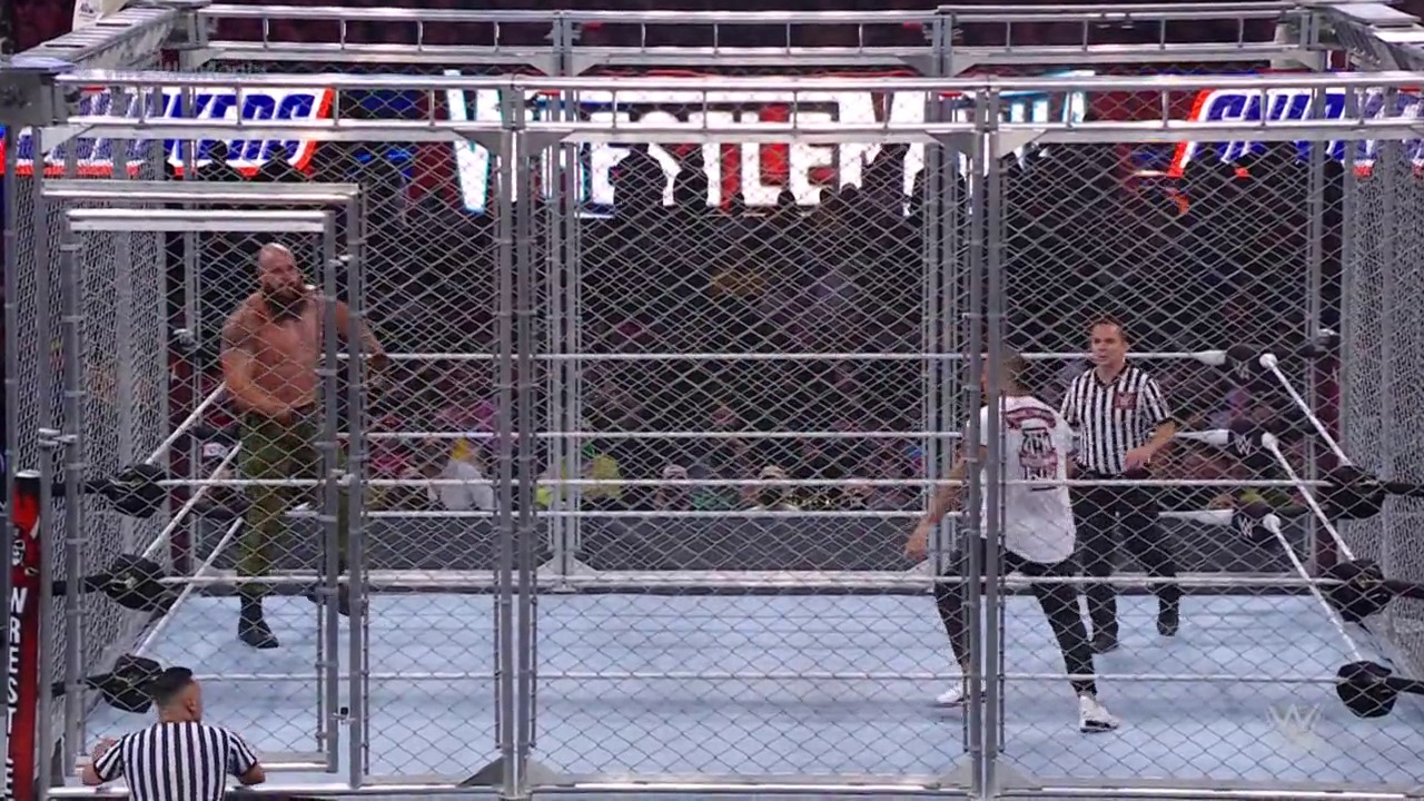 A frame from WrestleMania 37 (2021) at 1:48:07. A steel cage match at WrestleMania 37 features two wrestlers competing inside a large chain-link enclosed ring, with a referee monitoring the action and the crowd visible in the background.