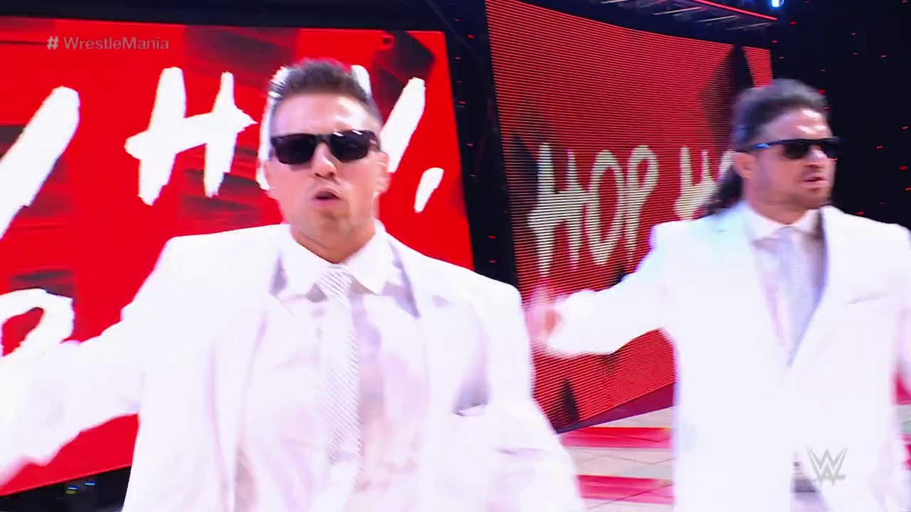 A frame from WrestleMania 37 (2021) at 2:14:51. Two wrestlers in white suits and sunglasses stand in the wrestling ring during WrestleMania 37, with red LED screens displaying graphics in the background.