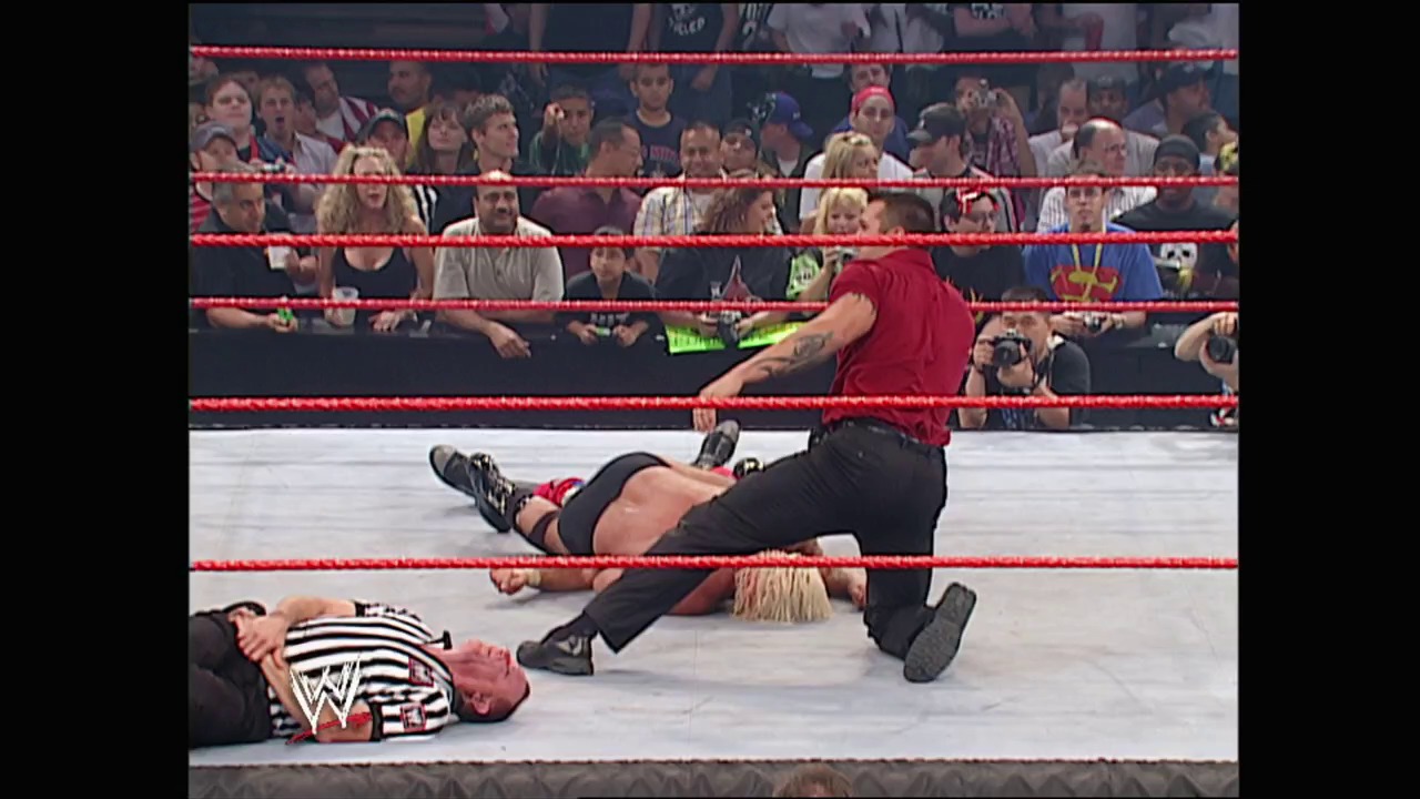 A frame from WWE Bad Blood (2003) at 1:48:11. Two WWE wrestlers are in action inside the red-roped ring, with one standing over another who is lying on the mat. The crowd can be seen in the background behind the ropes, and the WWE logo is visible on the wrestler's clothing.