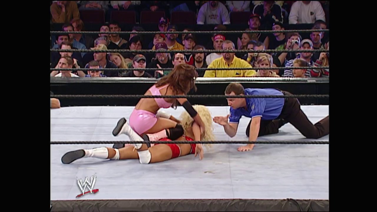 A frame from WWE No Mercy (2002) at 24:39. A WWE wrestling match in progress, with one wrestler in pink pinning an opponent in red and white on the mat while a referee in blue oversees the action. The crowd fills the arena stands in the background, with the WWE logo visible in the corner.