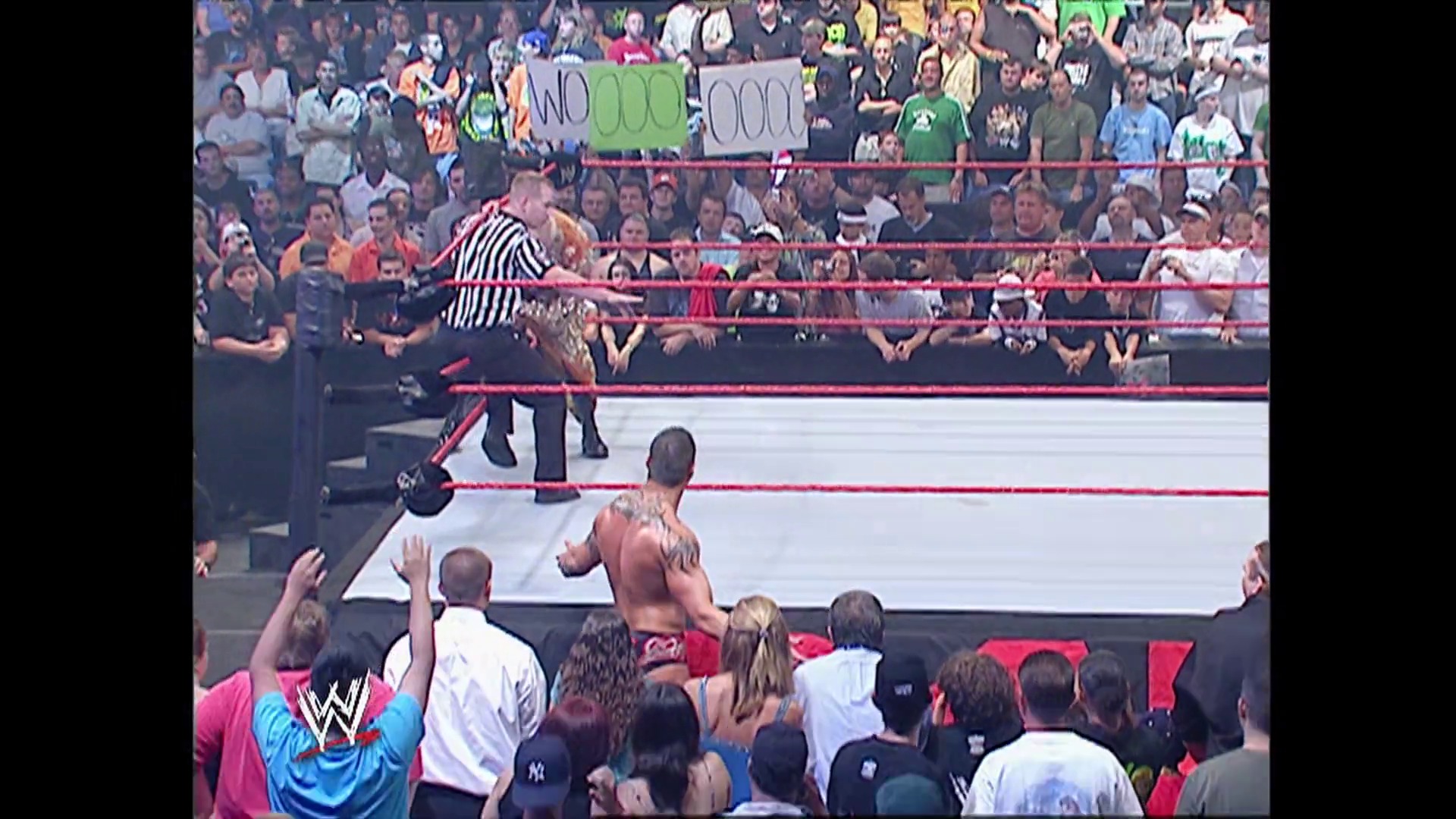A frame from WWE Raw (August 21, 2006) at 1:05:13. A WWE Raw match in progress inside a red-roped ring with a referee in striped shirt officiating. A shirtless wrestler engages with opponents while a packed crowd watches from the arena floor and stands, with the WWE logo visible in the lower left corner.