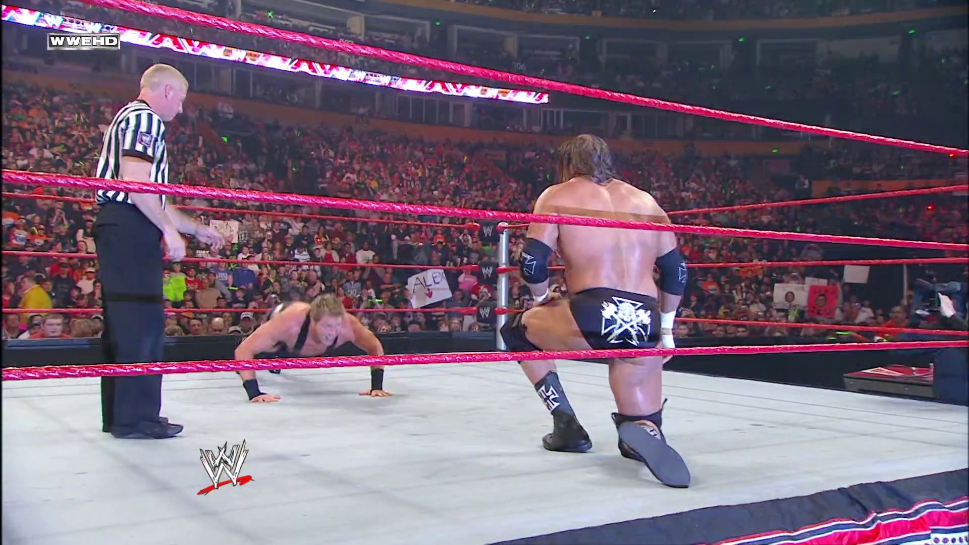 A frame from WWE Raw (February 1, 2010) at 27:59. A WWE Raw wrestling match in progress, featuring two shirtless wrestlers in the pink-roped ring with a referee in striped shirt overseeing. A packed crowd fills the arena in the background, with the WWE logo visible at the bottom of the screen.