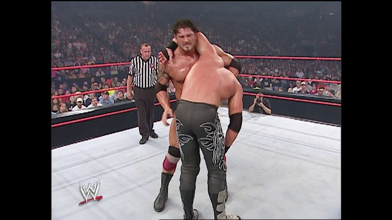 A frame from WWE Raw (July 12, 2004) at 14:28. A WWE wrestler carries another competitor over his shoulder in the ring during a match, with a referee in a striped shirt monitoring the action. The crowd fills the arena seats in the background, with red ropes framing the white wrestling ring.