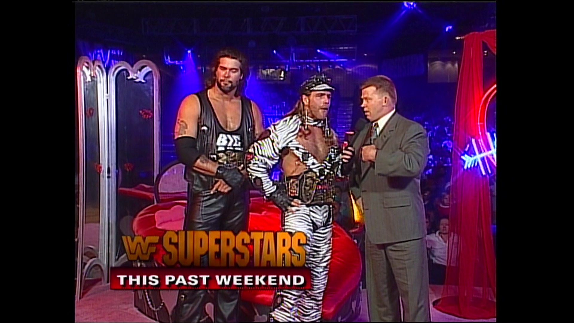 A frame from WWE Raw (October 10, 1994) at 16:27. # WWE Raw Visual Description

A WWE promoter in a gray suit interviews two wrestlers on the red-carpeted stage: one in black leather attire and another in a zebra-striped outfit with a crown. Neon signage and blue lighting frame the scene, with "WWE SUPERSTARS THIS PAST WEEKEND" displayed at the bottom.