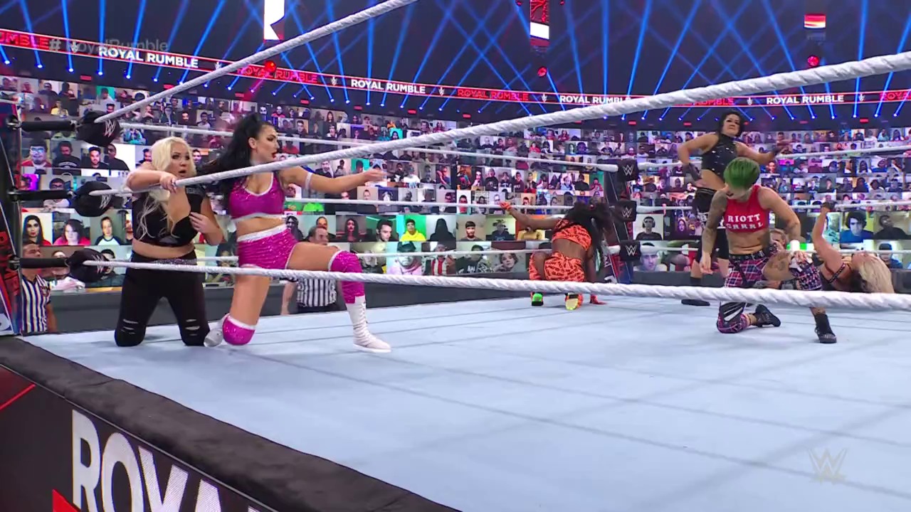 A frame from WWE: Royal Rumble (2021) at 1:05:24. WWE wrestlers compete in a Royal Rumble match inside a white-roped ring, with one wrestler in pink attire on the mat and others nearby. The arena features blue lighting, digital crowd screens, and prominent "Royal Rumble" branding around the venue.