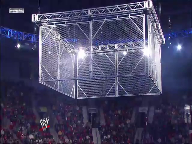 A frame from WWE SmackDown (January 15, 2010) at 1:11:47. A large steel cage structure is suspended above the wrestling ring, illuminated by bright spotlights in an arena filled with spectators. The WWE logo is visible in the lower right corner of the broadcast.