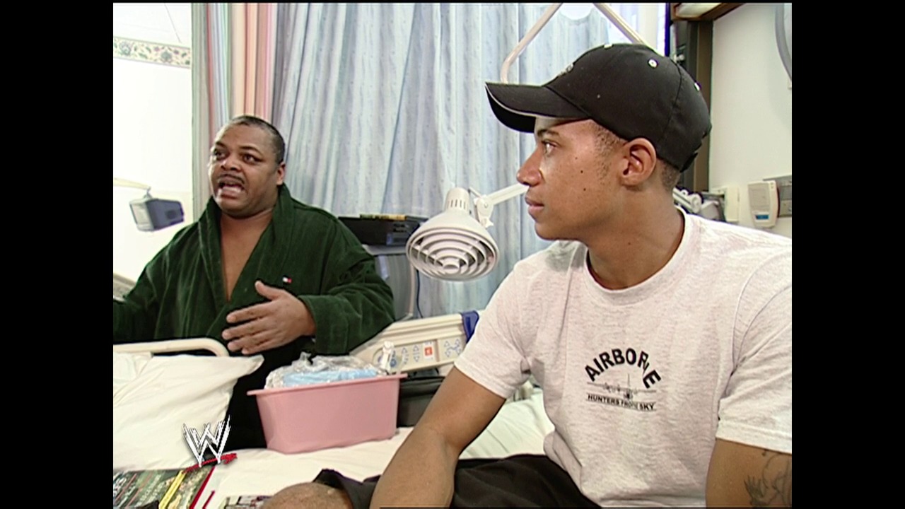 A frame from WWE SmackDown (January 29, 2004) at 48:56. Two men sit at a table in what appears to be a backstage area, with one wearing a green jacket on the left and another in an "Airborne" t-shirt and black cap on the right. A pink container and WWE graphics are visible on the table, with curtains and medical equipment in the background.