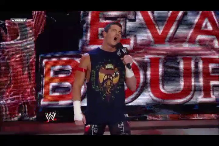 A frame from WWE Raw (July 11, 2011) at 43:46. A WWE wrestler stands in the ring holding a microphone, wearing a black band t-shirt and wrist tape, with a large LED screen displaying event graphics behind him during a WWE Raw broadcast.