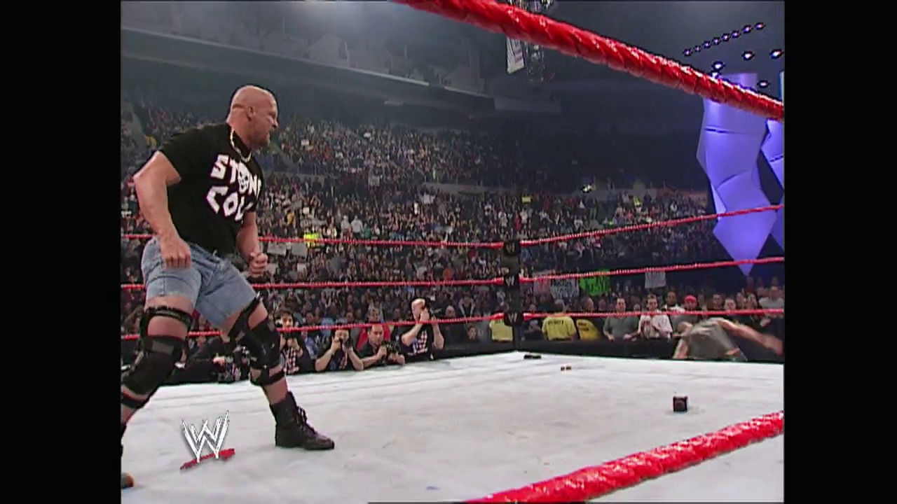 A frame from WWE Raw (March 3, 2003) at 1:26:44. A bald wrestler in a black "Stone Cold" shirt and jeans stands in a WWE ring with red ropes, preparing for action while a packed crowd watches from the arena stands. The WWE logo is visible on the mat, with blue lighting illuminating the venue.